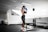 A person wearing athletic wear and a cap is in a gym setting, practicing boxing with blue gloves. The gym is well-lit and features fitness equipment such as dumbbells and benches. The person appears focused and is positioned in front of a mirrored wall.