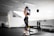 A person wearing athletic wear and a cap is in a gym setting, practicing boxing with blue gloves. The gym is well-lit and features fitness equipment such as dumbbells and benches. The person appears focused and is positioned in front of a mirrored wall.