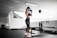 A person wearing athletic wear and a cap is in a gym setting, practicing boxing with blue gloves. The gym is well-lit and features fitness equipment such as dumbbells and benches. The person appears focused and is positioned in front of a mirrored wall.