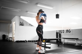 A person wearing athletic wear and a cap is in a gym setting, practicing boxing with blue gloves. The gym is well-lit and features fitness equipment such as dumbbells and benches. The person appears focused and is positioned in front of a mirrored wall.