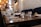 A dining area with wooden tables set with plates, upside-down glasses, and utensils. The seating includes plush, blue upholstered couches against a textured wall. There are warm pendant lights hanging from the ceiling, creating a cozy and inviting atmosphere. The background shows shelves and possibly a counter, hinting at a kitchen or serving area.