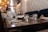 A dining area with wooden tables set with plates, upside-down glasses, and utensils. The seating includes plush, blue upholstered couches against a textured wall. There are warm pendant lights hanging from the ceiling, creating a cozy and inviting atmosphere. The background shows shelves and possibly a counter, hinting at a kitchen or serving area.