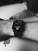A black and white image showing a person's wrist wearing a smart watch with a detailed screen display. The watch screen displays a graph with a time indicator and an instruction to 'Tap for Details'. The wrist is resting on a closed laptop, identifiable by a partially visible Apple logo.