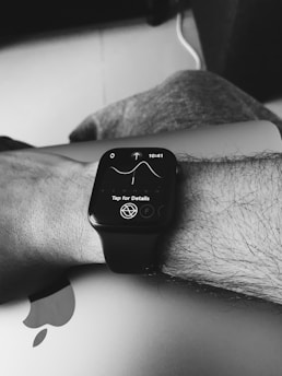 A black and white image showing a person's wrist wearing a smart watch with a detailed screen display. The watch screen displays a graph with a time indicator and an instruction to 'Tap for Details'. The wrist is resting on a closed laptop, identifiable by a partially visible Apple logo.