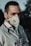 A person wearing a protective face mask and a stethoscope around their neck stands outdoors. The individual is dressed in a light-colored medical coat, suggesting they are a healthcare professional. The background is out of focus, with dark and green tones.