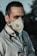 A person wearing a protective face mask and a stethoscope around their neck stands outdoors. The individual is dressed in a light-colored medical coat, suggesting they are a healthcare professional. The background is out of focus, with dark and green tones.