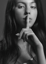 A black and white portrait of a woman with long hair holding her index finger to her lips in a gesture of silence. Her gaze is directed at the camera, creating an enigmatic and contemplative mood.