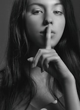 A black and white portrait of a woman with long hair holding her index finger to her lips in a gesture of silence. Her gaze is directed at the camera, creating an enigmatic and contemplative mood.