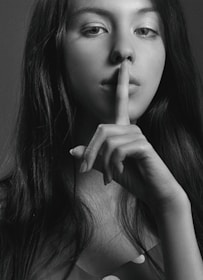 A black and white portrait of a woman with long hair holding her index finger to her lips in a gesture of silence. Her gaze is directed at the camera, creating an enigmatic and contemplative mood.