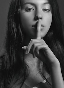 A black and white portrait of a woman with long hair holding her index finger to her lips in a gesture of silence. Her gaze is directed at the camera, creating an enigmatic and contemplative mood.