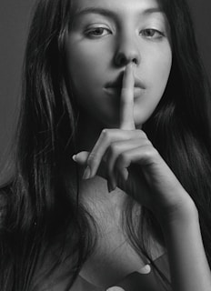 A black and white portrait of a woman with long hair holding her index finger to her lips in a gesture of silence. Her gaze is directed at the camera, creating an enigmatic and contemplative mood.
