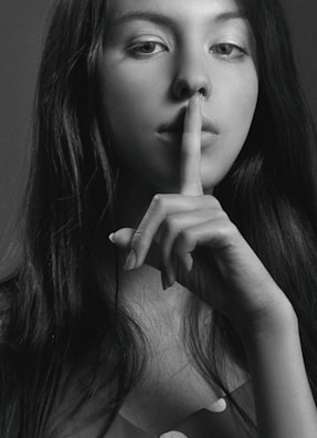A black and white portrait of a woman with long hair holding her index finger to her lips in a gesture of silence. Her gaze is directed at the camera, creating an enigmatic and contemplative mood.