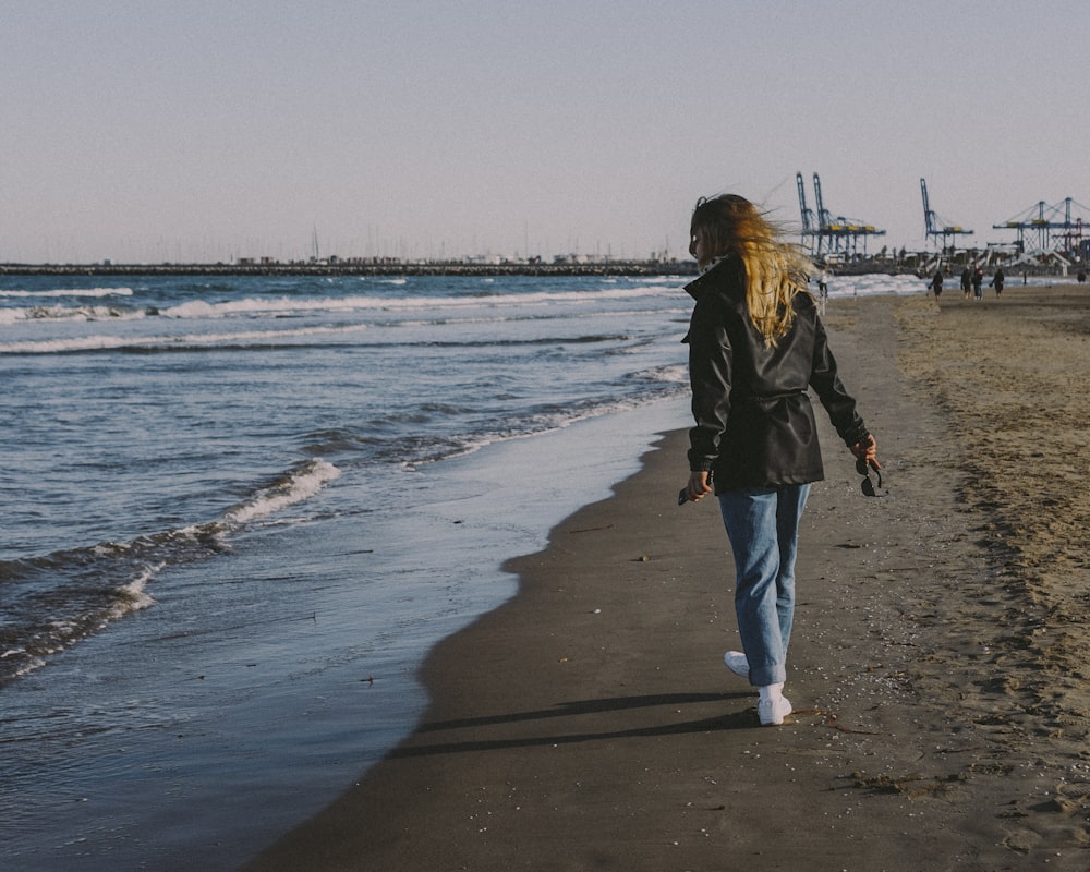 昼間のビーチを歩く黒いジャケットとブルーデニムジーンズの女性