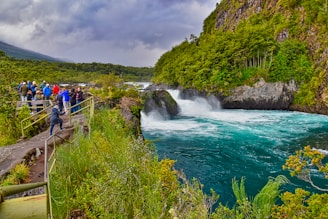 A group of people stand on a viewing platform overlooking a vibrant turquoise river that flows through rocky formations, surrounded by lush green trees. The sky is overcast, adding a dramatic backdrop to the natural scene. The river is turbulent, creating white water as it rushes through the landscape.