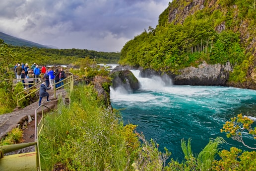A group of people stand on a viewing platform overlooking a vibrant turquoise river that flows through rocky formations, surrounded by lush green trees. The sky is overcast, adding a dramatic backdrop to the natural scene. The river is turbulent, creating white water as it rushes through the landscape.