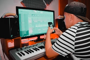 A person wearing a striped shirt and cap is using a digital audio workstation on a computer. They are also holding a smartphone. There is a keyboard, speakers, and soundproofing in the background, suggesting a music studio setting.