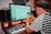 A person wearing a striped shirt and cap is using a digital audio workstation on a computer. They are also holding a smartphone. There is a keyboard, speakers, and soundproofing in the background, suggesting a music studio setting.