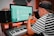 A person wearing a striped shirt and cap is using a digital audio workstation on a computer. They are also holding a smartphone. There is a keyboard, speakers, and soundproofing in the background, suggesting a music studio setting.
