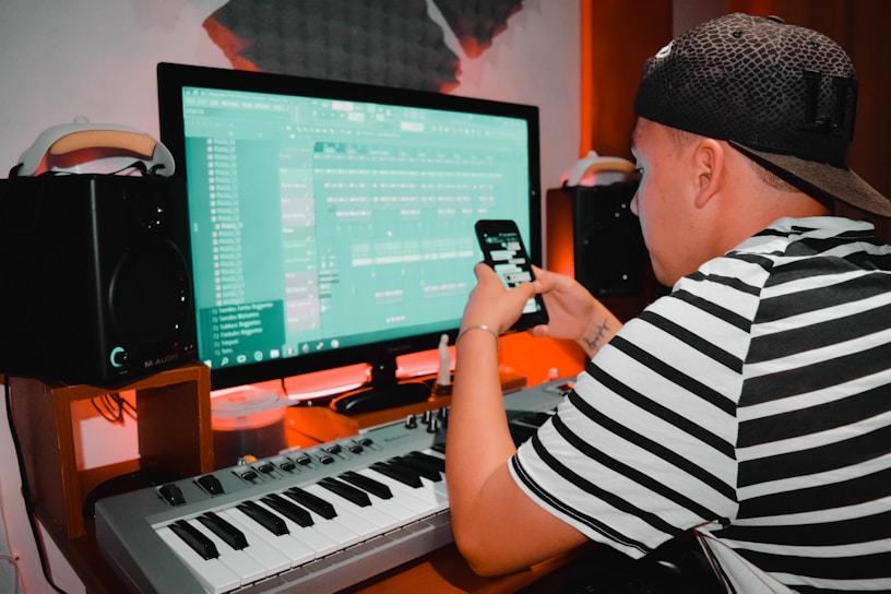 A person wearing a striped shirt and cap is using a digital audio workstation on a computer. They are also holding a smartphone. There is a keyboard, speakers, and soundproofing in the background, suggesting a music studio setting.