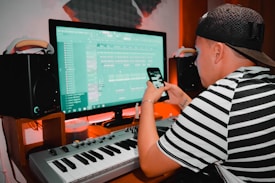 A person wearing a striped shirt and cap is using a digital audio workstation on a computer. They are also holding a smartphone. There is a keyboard, speakers, and soundproofing in the background, suggesting a music studio setting.