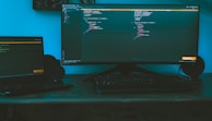 A dimly lit workspace featuring a large curved monitor displaying code, accompanied by a laptop to the side on a wooden desk. The ambient lighting casts a bluish hue, creating a focused and immersive environment. A keyboard is placed in front of the monitors, and speakers flank the large screen.