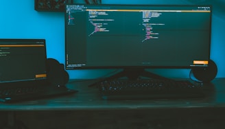 A dimly lit workspace featuring a large curved monitor displaying code, accompanied by a laptop to the side on a wooden desk. The ambient lighting casts a bluish hue, creating a focused and immersive environment. A keyboard is placed in front of the monitors, and speakers flank the large screen.