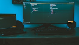 A dimly lit workspace featuring a large curved monitor displaying code, accompanied by a laptop to the side on a wooden desk. The ambient lighting casts a bluish hue, creating a focused and immersive environment. A keyboard is placed in front of the monitors, and speakers flank the large screen.
