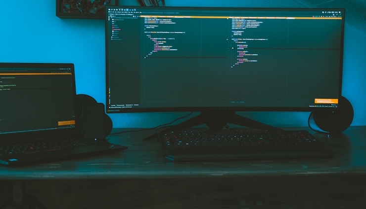 A dimly lit workspace featuring a large curved monitor displaying code, accompanied by a laptop to the side on a wooden desk. The ambient lighting casts a bluish hue, creating a focused and immersive environment. A keyboard is placed in front of the monitors, and speakers flank the large screen.