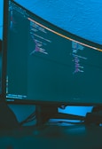 A large curved monitor displays a dark-themed code editor, featuring lines of code written in a structured format. The background is a textured ceiling with soft blue lighting creating a calm ambiance.