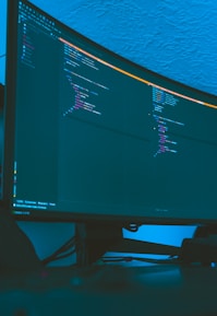 A large curved monitor displays a dark-themed code editor, featuring lines of code written in a structured format. The background is a textured ceiling with soft blue lighting creating a calm ambiance.