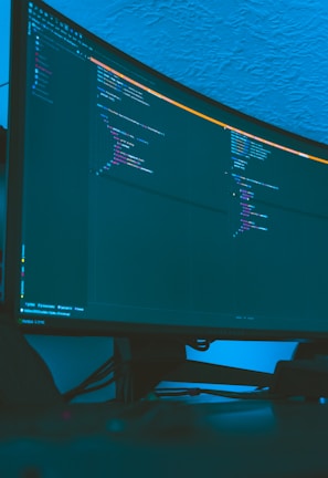 A large curved monitor displays a dark-themed code editor, featuring lines of code written in a structured format. The background is a textured ceiling with soft blue lighting creating a calm ambiance.