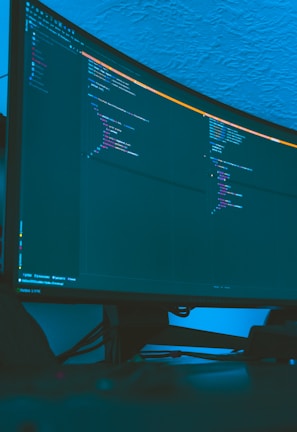 A large curved monitor displays a dark-themed code editor, featuring lines of code written in a structured format. The background is a textured ceiling with soft blue lighting creating a calm ambiance.