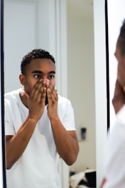 A person stands in front of a mirror with both hands covering the lower part of their face, appearing surprised or contemplative. The individual is wearing a white shirt and is indoors in a well-lit room.