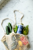 A mesh bag rests on a marble surface, filled with fresh produce and a jar of vegan tzatziki sauce. Inside the bag are cucumbers, greens, a lemon, and a red bell pepper. The jar has a blue label with the brand name 'FABALISH'.