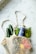 A mesh bag rests on a marble surface, filled with fresh produce and a jar of vegan tzatziki sauce. Inside the bag are cucumbers, greens, a lemon, and a red bell pepper. The jar has a blue label with the brand name 'FABALISH'.