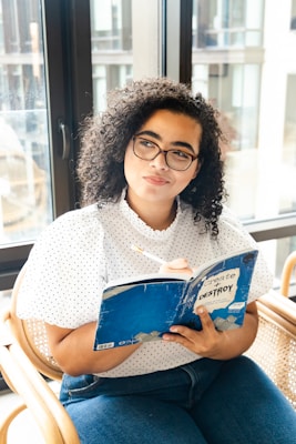 A person with curly hair wearing glasses, a white polka dot blouse, and jeans is seated on a chair near a large window, holding a notebook with a blue cover titled 'Create or Destroy' and a pencil in hand.