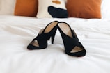 A pair of stylish black heeled sandals are placed on a neatly made white bed, with two brown cushions and a decorative pillow in the background.