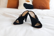 A pair of stylish black heeled sandals are placed on a neatly made white bed, with two brown cushions and a decorative pillow in the background.
