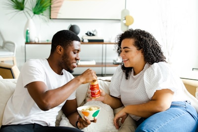 A man and a woman are sitting on a couch, enjoying a snack together. The man, wearing a white t-shirt, is holding a bowl and pouring sauce from a bottle, while the woman in a white polka dot top and jeans watches and smiles. The setting appears to be a cozy and modern living room with a decorative plant and some abstract artwork in the background.