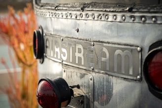 Close-up of a vintage Airstream trailer's rear end, displaying its metal nameplate and circular red taillight against a blurred background of nature.