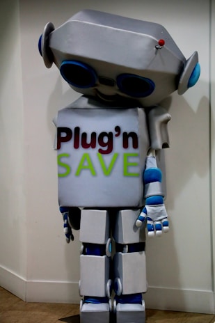A humanoid robot costume with a metallic gray and blue color scheme, boxy limbs, and a large rectangular torso. The robot has a large head with circular antenna on top and large, round blue eyes. The front of the torso features the text 'Plug'n SAVE' in bold letters.