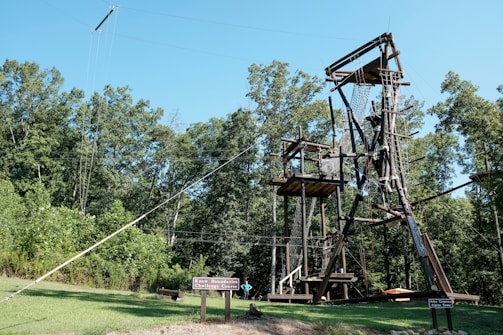 A wooden adventure course structure stands amidst lush green trees. It includes platforms, ladders, and netting designed for climbing and exploring. The sky is clear and blue, creating a feeling of outdoor adventure. A small sign reads 'Challenge Course'.