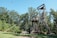 A wooden adventure course structure stands amidst lush green trees. It includes platforms, ladders, and netting designed for climbing and exploring. The sky is clear and blue, creating a feeling of outdoor adventure. A small sign reads 'Challenge Course'.