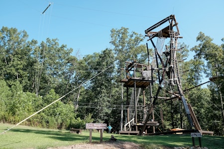 A wooden adventure course structure stands amidst lush green trees. It includes platforms, ladders, and netting designed for climbing and exploring. The sky is clear and blue, creating a feeling of outdoor adventure. A small sign reads 'Challenge Course'.