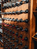 A wooden wall-mounted wine rack displays numerous bottles stored horizontally, each held securely by metal holders. The warm tones of the wood contrast with the dark colors of the wine bottles, creating an elegant and orderly arrangement.