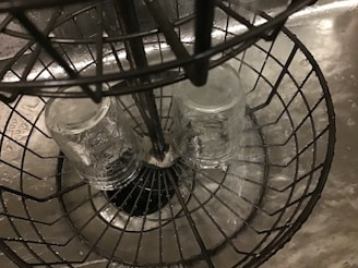 Two clear glasses are placed upside down in a wire rack within a kitchen sink. Water droplets are visible on the surfaces of the glasses and the sink.