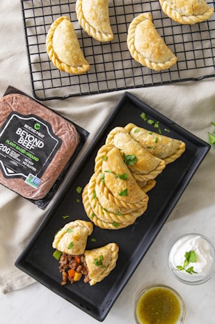 A set of baked empanadas displayed on a black cooling rack and a rectangular black plate, with a filling of plant-based meat and vegetables visible in one cut-open empanada. Adjacent to them is a package of plant-based ground meat labeled 'Beyond Beef' and two small bowls containing sour cream garnished with cilantro and a green salsa.
