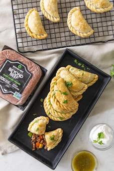 A set of baked empanadas displayed on a black cooling rack and a rectangular black plate, with a filling of plant-based meat and vegetables visible in one cut-open empanada. Adjacent to them is a package of plant-based ground meat labeled 'Beyond Beef' and two small bowls containing sour cream garnished with cilantro and a green salsa.