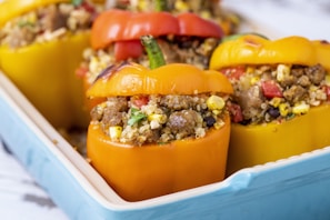 Stuffed bell peppers filled with a mixture of quinoa, ground meat, corn, black beans, and diced vegetables are arranged in a blue baking dish. The peppers are vibrant in color, featuring red, yellow, and orange hues.