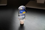 A small, transparent hand sanitizer bottle with a blue flip cap is placed on a dark surface. The bottle features a blue label with white text and a clip attached to the top.